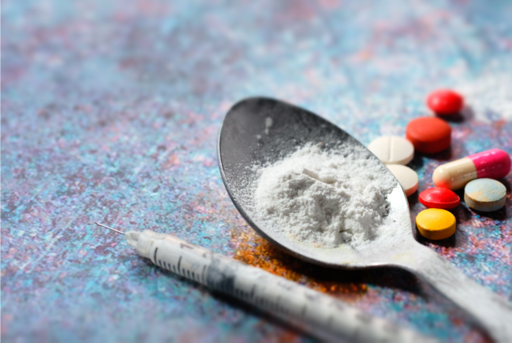 Imagem mostrando pó branco sobre uma colher metálica, seringa descartável e comprimidos coloridos ao fundo, representando riscos associados ao uso de drogas injetáveis, dependência química e problemas de saúde pública relacionados a substâncias ilícitas.